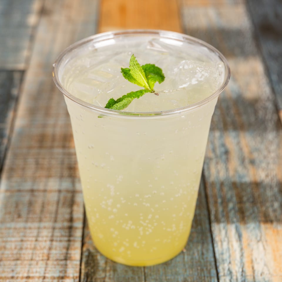 Bubbly light yellow drink with ice and mint in a plastic cup on a wooden table.
