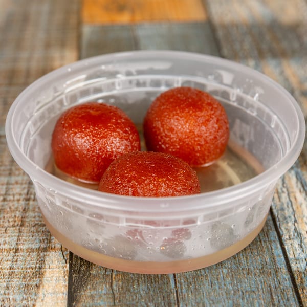 Three Gulab Jamun sweets in syrup inside a clear plastic container on a wooden table.