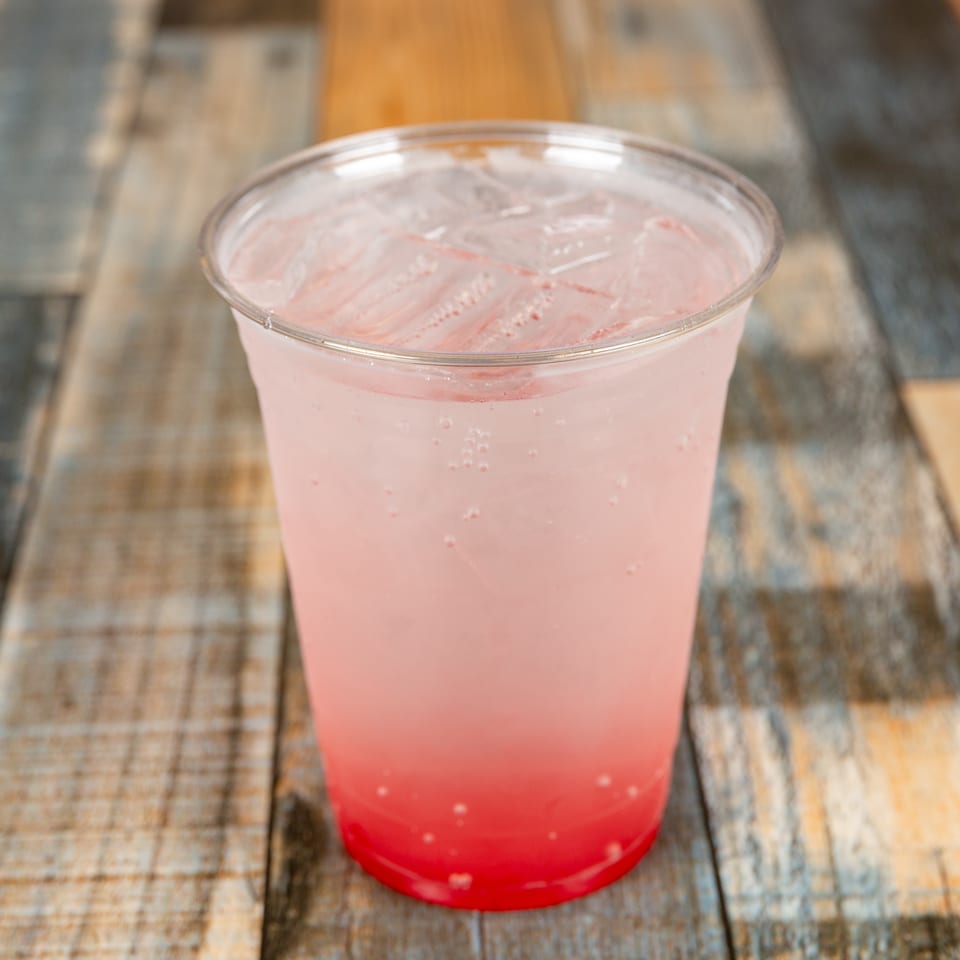 Pink fizzy drink with ice in a clear plastic cup on a rustic wooden table.