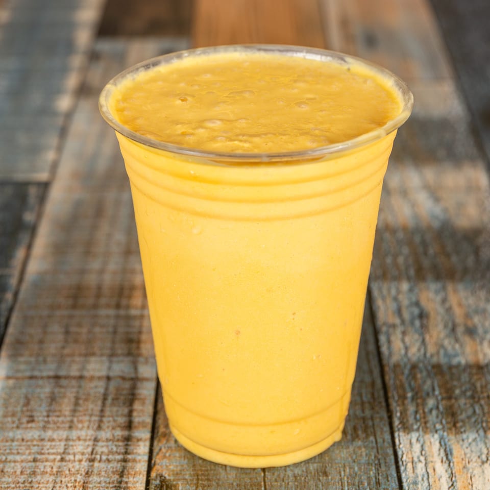 A vibrant yellow smoothie in a plastic cup on a distressed wooden surface.