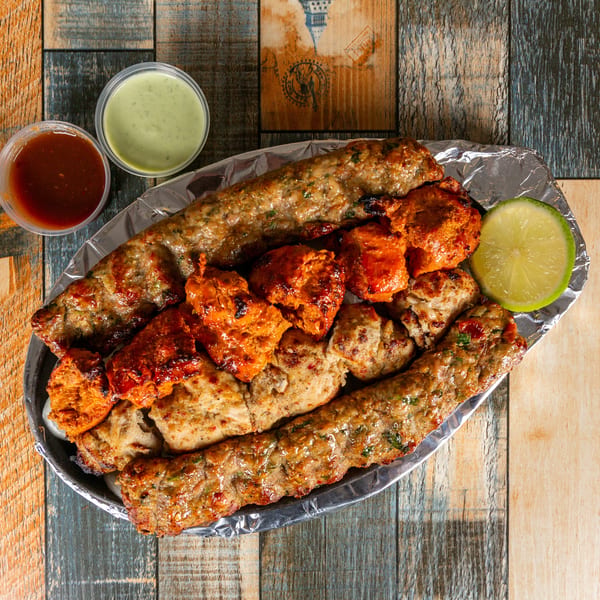 Platter of grilled kebabs and chicken tikka with red and green sauces, and a lime slice on wood.