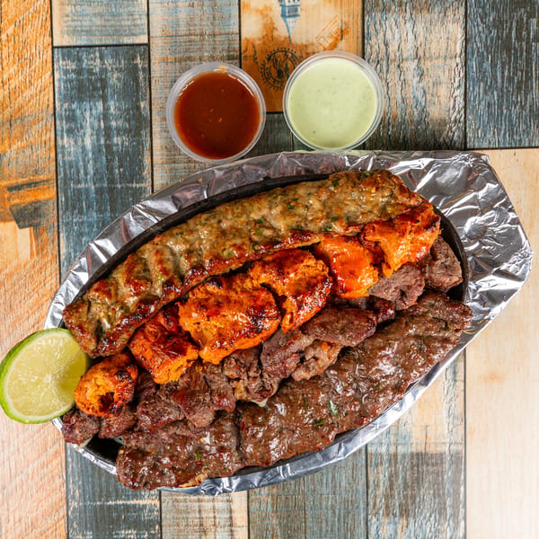 Platter of grilled kebabs, chicken tikka, and beef with dipping sauces and a lime wedge on a wooden table.